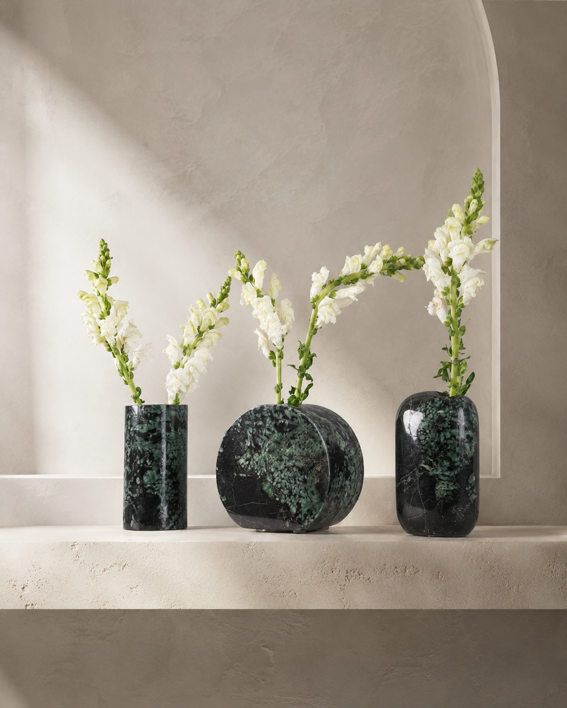 Trio de vasos de mármore verde-escuro em formatos geométricos sobre prateleira neutra, com hastes de flores brancas em composição minimalista.
